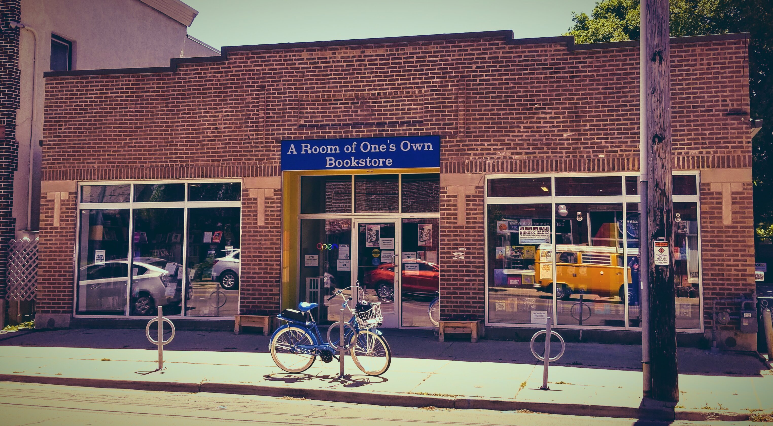 Exterior of A Room of One's Own Bookstore, a brick storefront with a blue sign above the entrance. A blue bicycle with a basket is parked at a bike rack in front. Large display windows flank the central entrance, with cars visible through the windows and additional storefronts visible on either side.