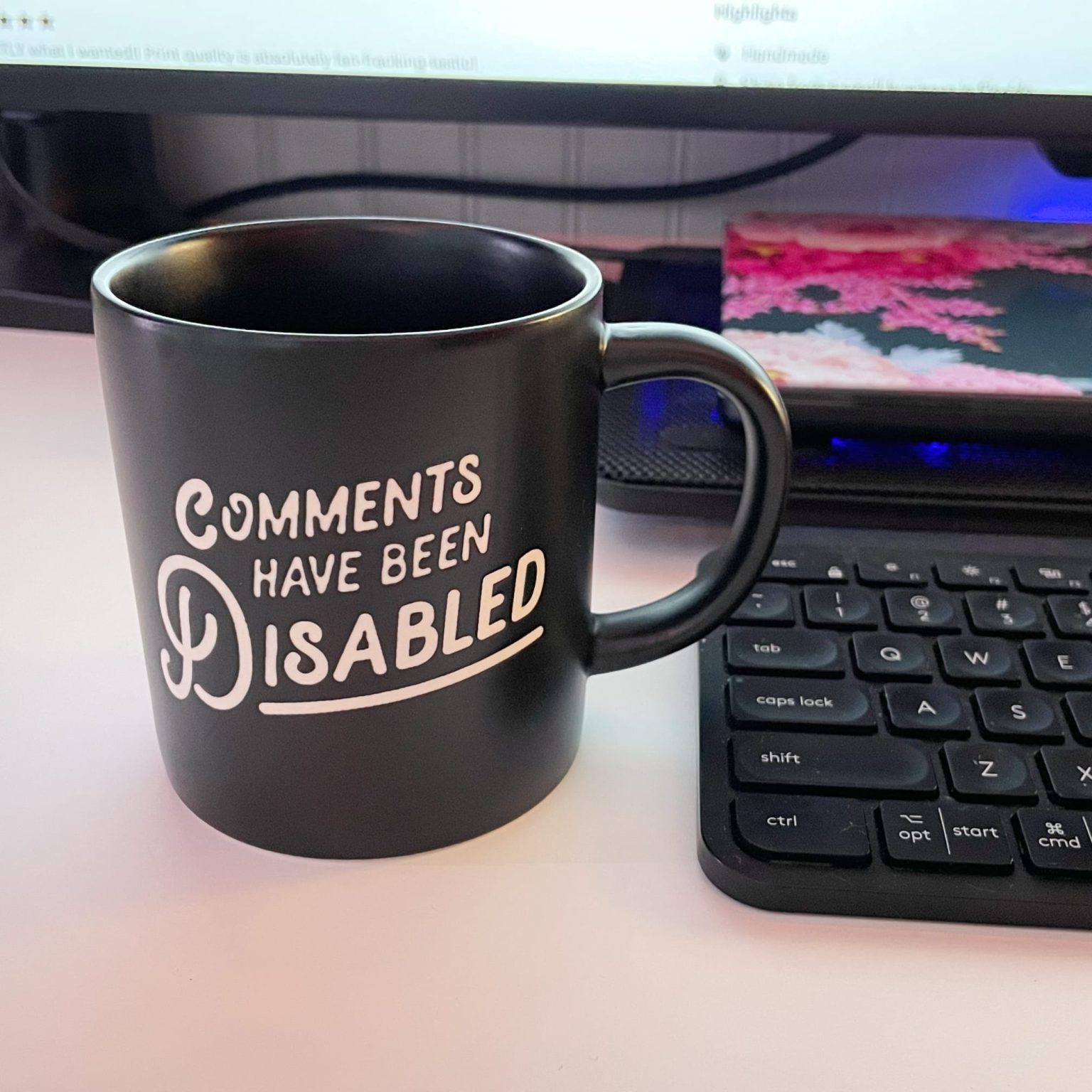 Black coffee mug on a desk displaying white text that reads 'COMMENTS HAVE BEEN DISABLED' in a playful handwritten font. The mug sits in front of a laptop keyboard with a monitor visible in the background, creating a typical home office workspace setting.