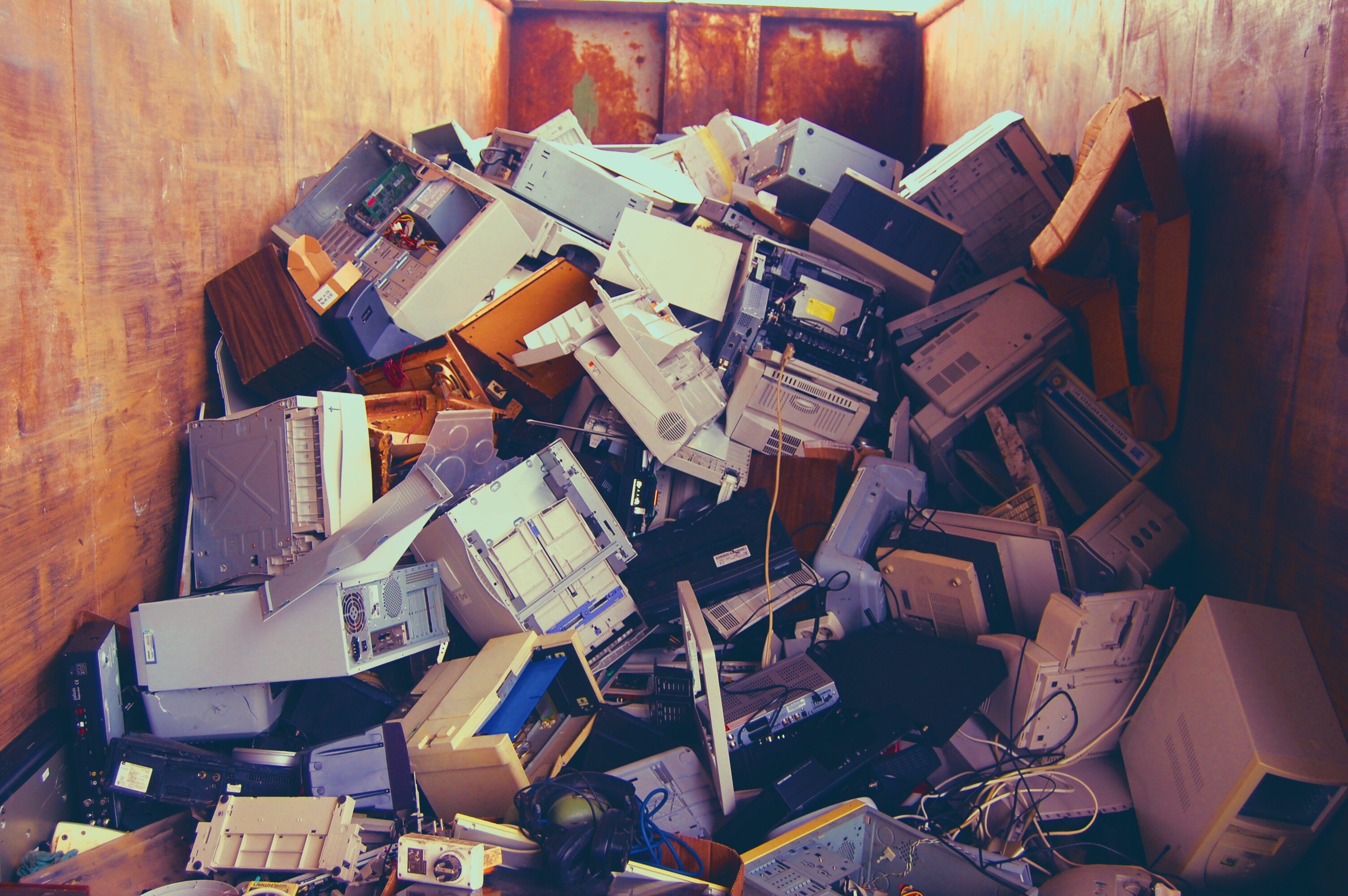 A large metal dumpster filled with discarded electronic equipment, including old desktop computer towers, printers, monitors, keyboards, and tangled cables piled chaotically inside. The interior walls of the dumpster are rust-stained, and the electronics are outdated and broken, electronic waste and bulk disposal.