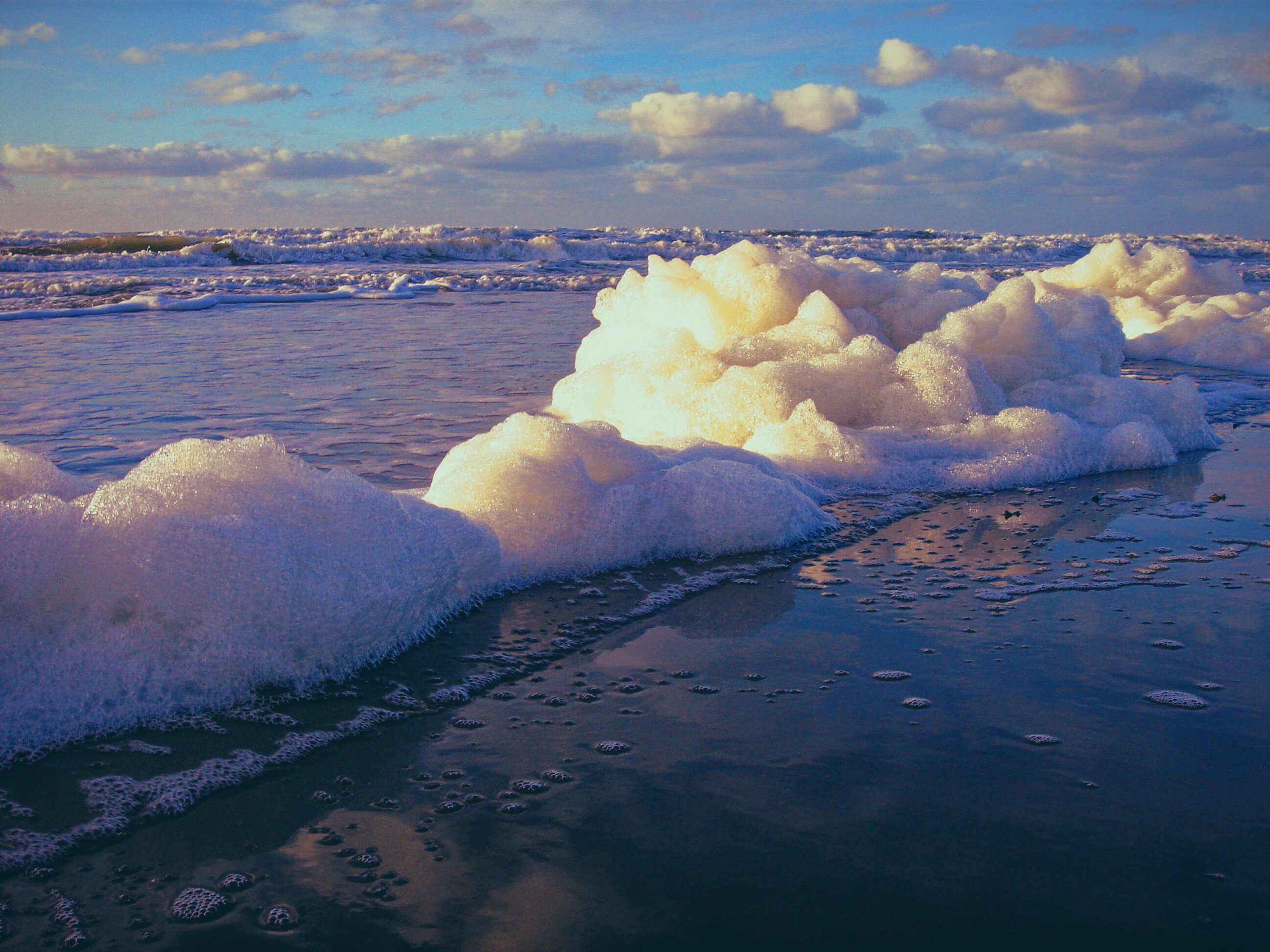 Mounds of sea foam pile up on a wet sandy beach at golden hour, glowing warm amber and white where sunlight catches their peaks. Shallow water reflects the purple-blue sky behind them, and rolling waves break in the background beneath a partly cloudy horizon.