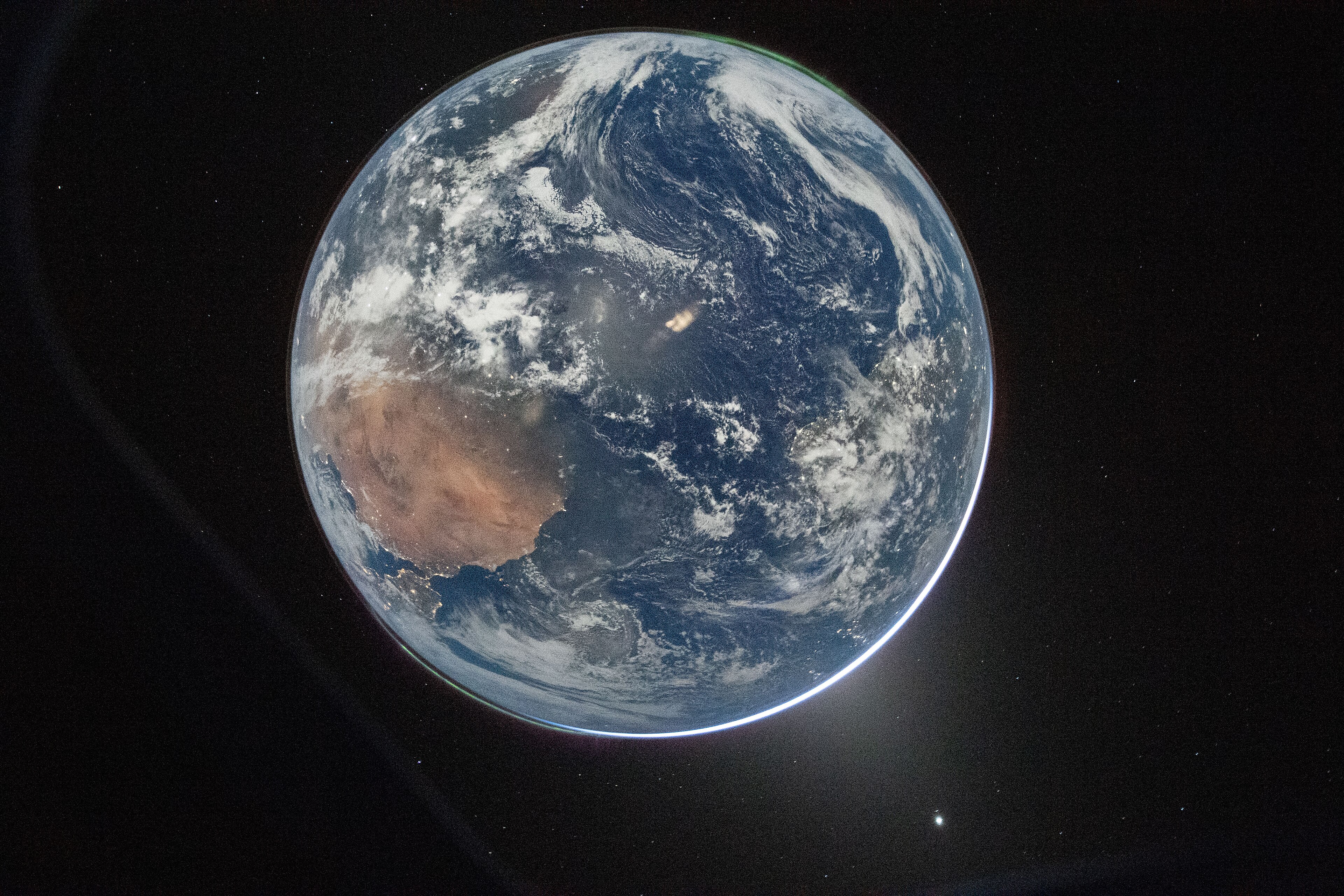 A photograph of Earth from deep space, slightly off-centre and partially in shadow, with a thin luminous blue atmospheric halo visible along the sunlit edge. Australia's arid interior is visible as a rust-coloured landmass on the left side. Swirling cloud systems cover much of the Pacific Ocean to the right. Stars are faintly visible in the black background, and a bright lens flare appears near the lower right.