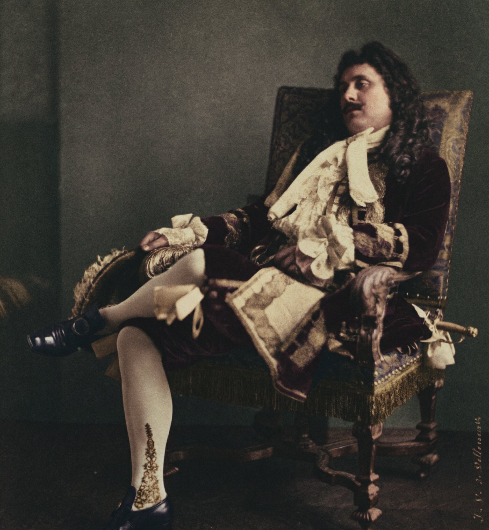 A hand-colored photograph of a male stage actor in 17th-century French theatrical costume, reclining languidly in an ornate gilded armchair. He wears a deep burgundy velvet coat with elaborate lace cravat and cuffs, white stockings with gold embroidered detail, and black heeled shoes.