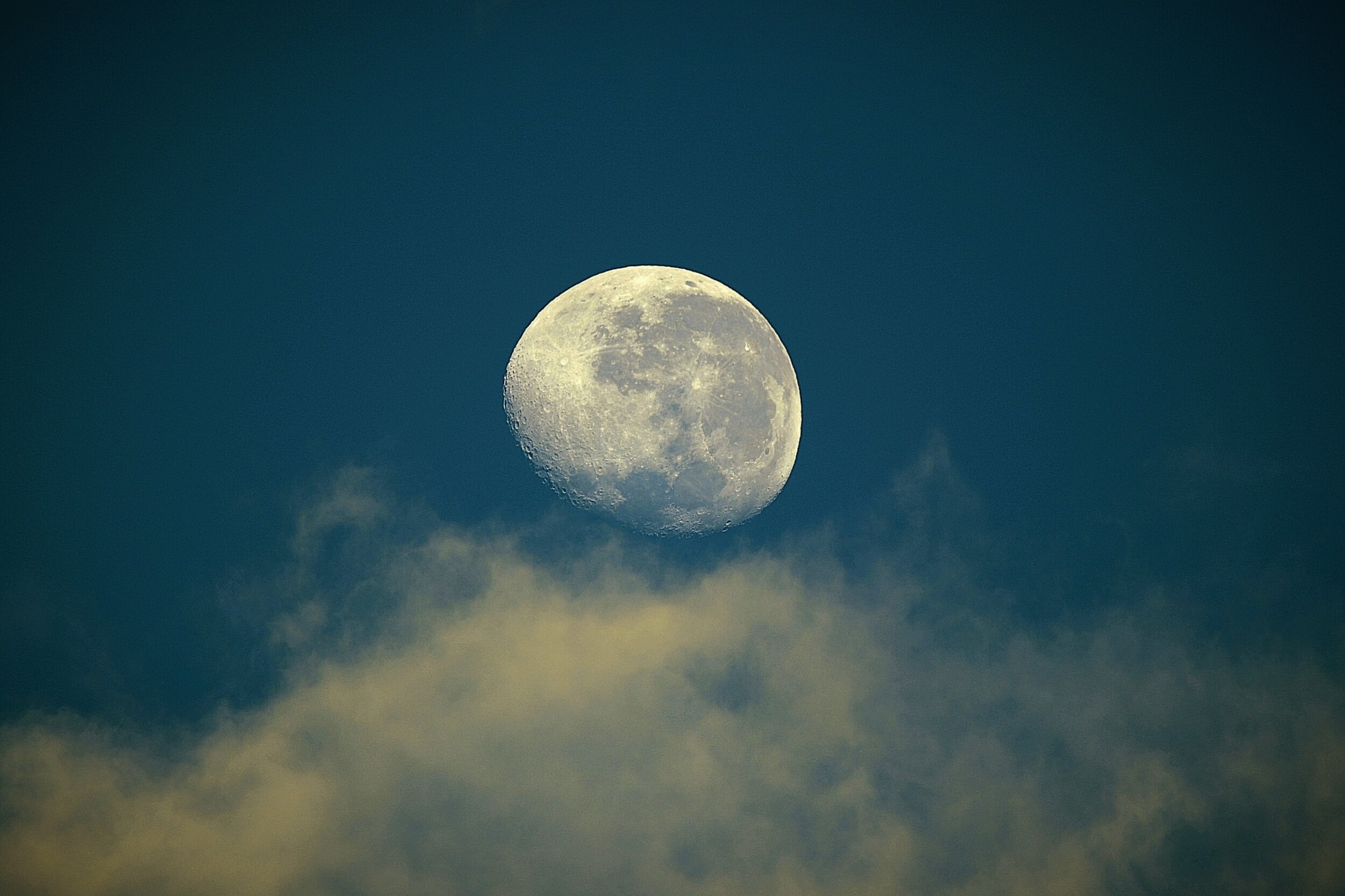 A waxing gibbous moon photographed against a deep teal night sky, rising above a bank of soft cumulus clouds. The lunar surface is sharply detailed, with visible craters and maria. The clouds in the foreground are lit from below with a warm golden tone, contrasting with the cool blue of the sky.