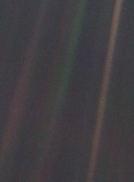 A grainy, dark photograph taken from deep space showing an almost featureless expanse of black and dark grey. A bright diagonal shaft of scattered sunlight crosses the right side of the frame. Near the centre of this beam, barely distinguishable from the surrounding darkness, is a tiny pale bluish-white speck — Earth, photographed from approximately 6 billion kilometres away by the Voyager 1 spacecraft in 1990.