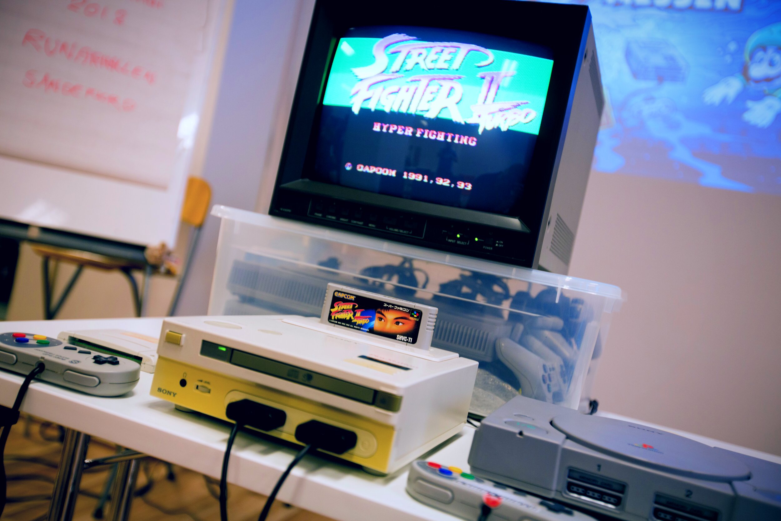 A retro gaming setup on a white table features a Nintendo Playstation prototype game console with a cartridge inserted, connected controllers, and a gray PlayStation nearby. Behind the consoles, a CRT monitor displays the Street Fighter II Turbo: Hyper Fighting title screen, while additional controllers are stored in a clear plastic bin. The softly blurred background includes a whiteboard and colourful game-themed artwork.