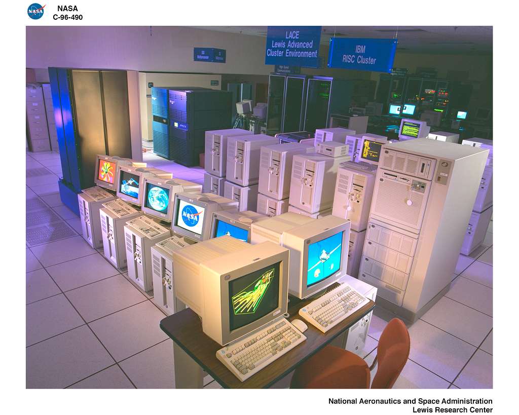 Interior view of a computer facility at NASA's Lewis Research Center showing rows of 1990s-era workstations and servers. The room features multiple beige computer towers and CRT monitors displaying various graphics including the NASA logo, Earth imagery, and scientific visualizations. Overhead signs identify the 'LACE Lewis Advanced Cluster Environment' and 'IBM RISC Cluster' systems.