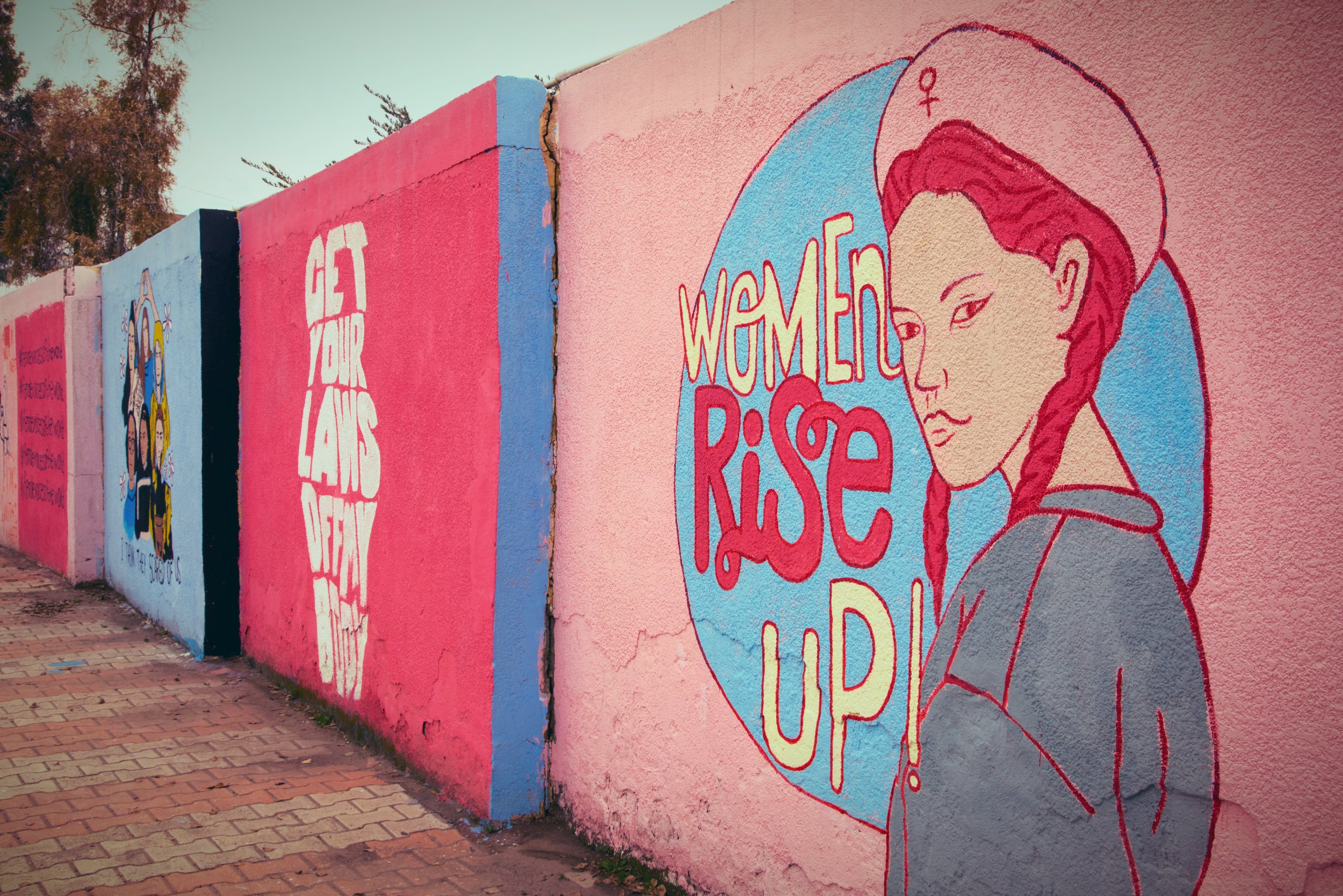 A series of feminist murals painted on a long outdoor wall in the Kurdistan Region of Iraq. The foreground mural on a pink background depicts a illustrated woman with a red braid wearing a beret bearing the Venus/female symbol, beside the text 'Women Rise Up!' in bold red lettering within a blue circle. Behind it, a pink panel reads 'Get Your Laws Off My Body' in white text. Further along the wall, a blue panel features a group illustration of diverse women. The wall runs alongside a brick-paved walkway, with trees visible in the background.