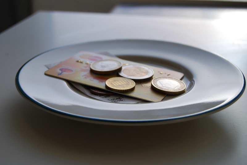 A white ceramic plate with a thin blue rim holds several euro coins of different denominations and a folded 10 euro banknote, arranged on a dark gray table or countertop with a blurred background.