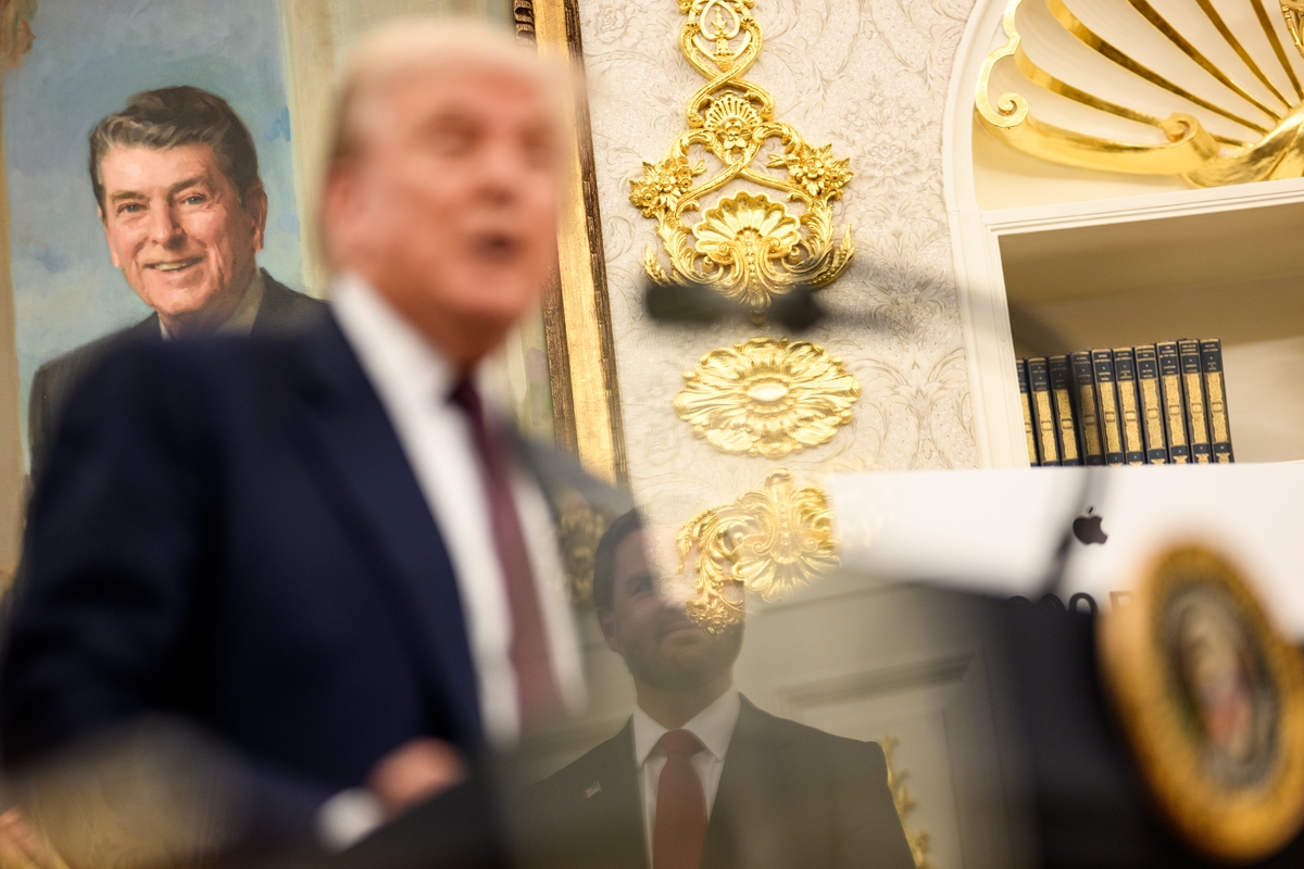 A blurred figure speaks at a podium bearing a presidential seal in an ornately decorated room with gold baroque wall moldings and white paneling. Behind them hangs a painted portrait of a smiling older man in a dark suit. A bookshelf with dark-spined volumes and an Apple laptop are visible to the right. A second figure in a suit is reflected in a glass surface in the foreground.