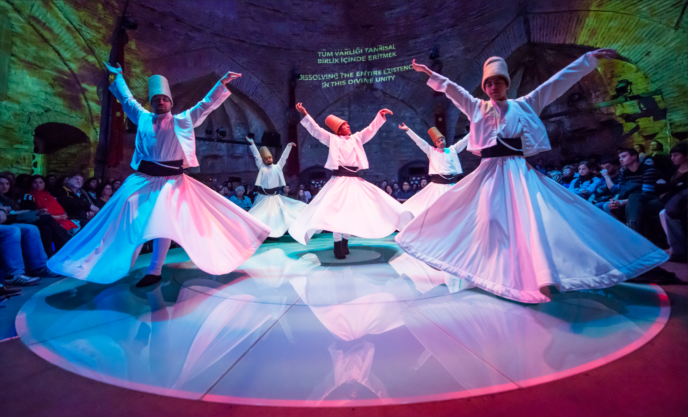 Whirling dervishes performing a traditional Sufi sema ceremony in a historic stone venue with arched architecture. Five performers in white robes and tall brown hats spin with arms outstretched, their wide skirts creating flowing circular patterns.