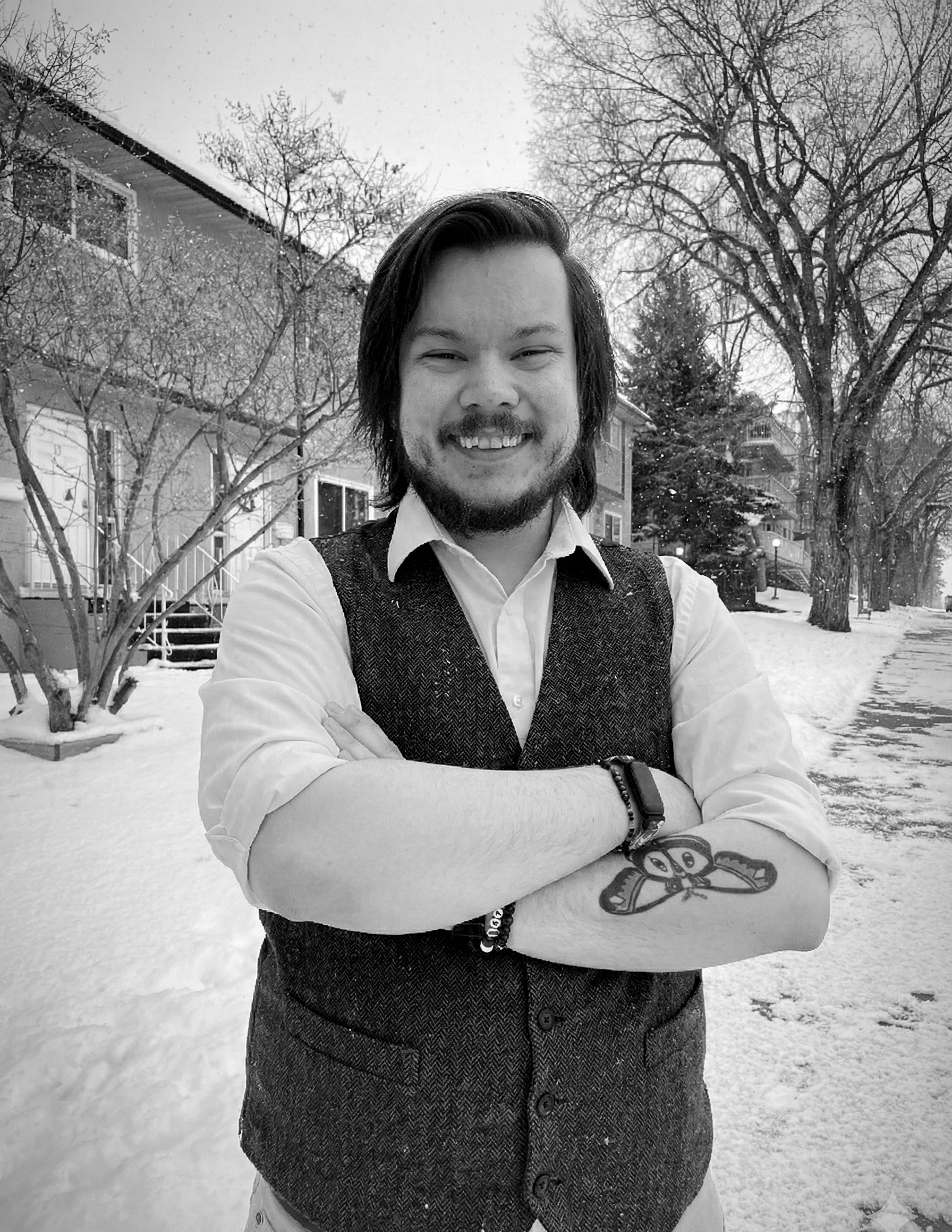 Black and white photograph of me, Brennan, with shoulder-length dark hair and a beard, smiling warmly at the camera on a snowy residential street. I'm wearing a white dress shirt with a dark herringbone tweed vest.