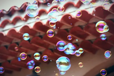 Dozens of soap bubbles float upward against a backdrop of terracotta roof tiles shot from below. The bubbles range in size from small to large and display iridescent colours: electric blue, magenta, gold, and green. All shimmering across their thin, transparent surfaces. Some bubbles reflect distorted glimpses of sky and surroundings. The roof tiles are rendered soft and out of focus in warm red-brown tones, creating contrast with the bright, jewel-like spheres in the foreground.