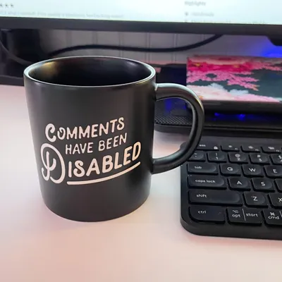 Black coffee mug on a desk displaying white text that reads 'COMMENTS HAVE BEEN DISABLED' in a playful handwritten font. The mug sits in front of a laptop keyboard with a monitor visible in the background, creating a typical home office workspace setting.