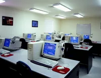 A bright computer lab with rows of desktop computers arranged on long tables. Each workstation features a bulky CRT monitor, tower computer, keyboard, mouse on a red mouse pad, and a headset hanging from the monitor. A whiteboard is mounted on the back wall, framed posters decorate the room, and fluorescent ceiling lights illuminate the clean, empty classroom.