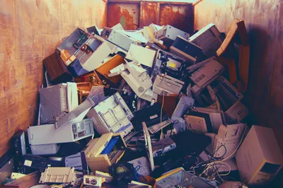 A large metal dumpster filled with discarded electronic equipment, including old desktop computer towers, printers, monitors, keyboards, and tangled cables piled chaotically inside. The interior walls of the dumpster are rust-stained, and the electronics are outdated and broken, electronic waste and bulk disposal.