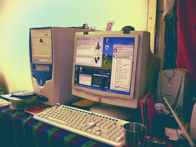A early-2000s home computer setup featuring a beige CRT monitor displaying Windows XP with MSN Messenger open, alongside a matching beige tower PC with a DVD drive and blue accent panel. The desk is covered with a striped cloth, and a beige keyboard sits in the foreground next to a metal cup. Small figurines and a photo are propped on top of the monitor. A speaker is visible to the right, and red curtains hang in the background. The photo has a warm, faded green-yellow tint 