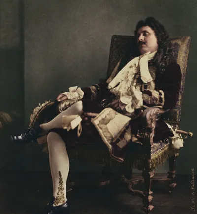 A hand-colored photograph of a male stage actor in 17th-century French theatrical costume, reclining languidly in an ornate gilded armchair. He wears a deep burgundy velvet coat with elaborate lace cravat and cuffs, white stockings with gold embroidered detail, and black heeled shoes.