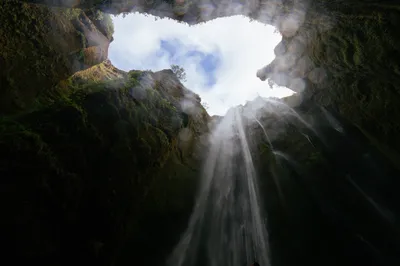 A dark pit with a waterfall cascading down steep moss-covered cliff walls, viewed from below, with mist rising through the narrow canyon opening to a cloudy sky above