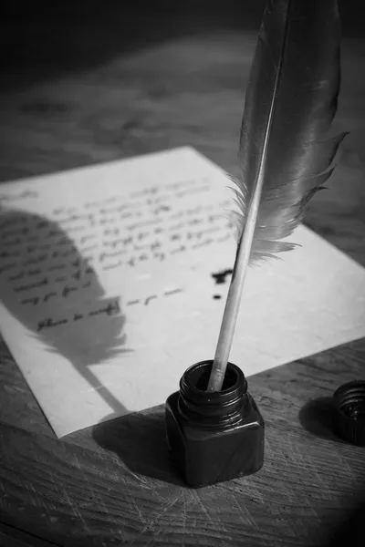 A quill pen rests upright in an open black ink bottle on a wooden table, with a handwritten letter on parchment paper lying blurred in the background.