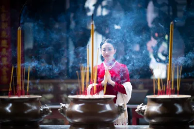 A young woman in traditional red and white Asian attire stands with hands pressed together in prayer position (anjali mudra) at a temple altar. She is centered among three bronze incense burners filled with burning incense sticks, with smoke rising around her. The atmospheric scene shows ornate temple architecture with decorative murals in the blurred background.
