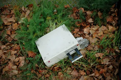 Weathered computer with visible dirt and moss growth abandoned on the ground among scattered brown autumn leaves, green grass patches, and foliage.