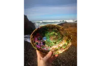 A hand holding an abalone shell up toward the camera on a rocky beach, displaying its iridescent interior of swirling greens, purples, pinks, and golds with characteristic respiratory holes visible on the left side. Crashing ocean waves and dark coastal rocks are visible in the blurred background under a partly cloudy sky.