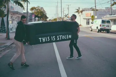 Two men carrying a dark green couch across a street in a tropical urban setting with palm trees. The couch has 'THIS IS STOLEN' written on it in large white hand-painted letters. Background shows commercial buildings, power lines, and vehicles on what appears to be a sunny day.