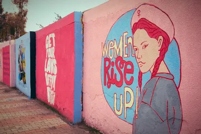 A series of feminist murals painted on a long outdoor wall in the Kurdistan Region of Iraq. The foreground mural on a pink background depicts a illustrated woman with a red braid wearing a beret bearing the Venus/female symbol, beside the text 'Women Rise Up!' in bold red lettering within a blue circle. Behind it, a pink panel reads 'Get Your Laws Off My Body' in white text. Further along the wall, a blue panel features a group illustration of diverse women. The wall runs alongside a brick-paved walkway, with trees visible in the background.