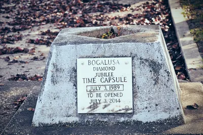 A weathered concrete time capsule monument sitting on a sidewalk surrounded by fallen leaves. A metal plaque on the front reads: 'Bogalusa Diamond Jubilee Time Capsule, July 3, 1989, To Be Opened July 3, 2014.' The top of the monument has cracked open, with small weeds growing through the gap.