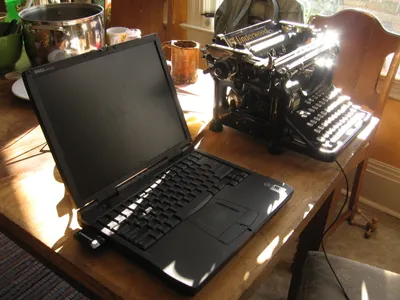 A black Dell laptop with blank screen sits open on a wooden desk next to a vintage black Underwood typewriter, bathed in natural sunlight streaming through a window. Behind them are a large stainless steel pot, amber-colored glassware, and a wooden chair. An evolution of writing tools.